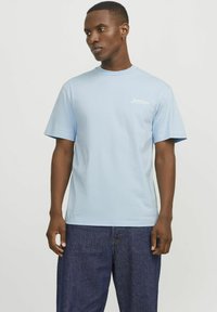 Camiseta de algodón de color azul claro con mangas cortas, cuello redondo y estampado de logo en la parte delantera. Ajuste holgado, combinada con jeans de mezclilla oscura.