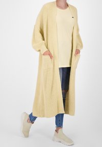 Długi dzianinowy kardigan w jasnym żółtym kolorze, z otwartym przodem, luźnymi rękawami i dwoma przednimi kieszeniami; noszony na pasującej żółtej bluzce i jeansach.