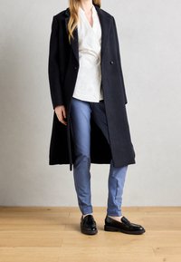 Frau steht auf einem Holzboden und trägt einen schwarzen langen Mantel, eine weiße Bluse, hellblaue Hosen und schwarze Lederloafers.