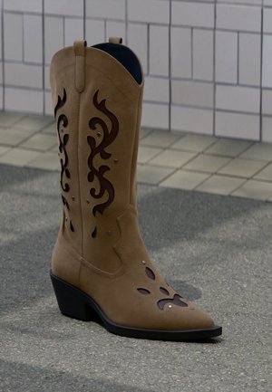 Botte de cowboy en daim beige avec des découpes décoratives marron foncé et des coutures, posée sur un trottoir devant un mur carrelé.