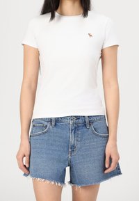 Camiseta blanca de manga corta con un pequeño logo marrón en el lado izquierdo del pecho, combinada con pantalones cortos de mezclilla azules con dobladillo deshilachado y bolsillos frontales.