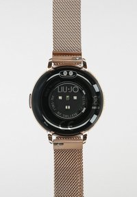 Smartwatch med en rund sort urskive, guld mesh-rem og cirkulær bagside med sensorer. Har mærket "LIU JO" og CE-mærkning.