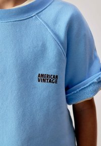 Enfant portant un sweat-shirt à manches courtes bleu clair avec des poignets retroussés et un petit texte "AMERICAN VINTAGE" sur la poitrine.