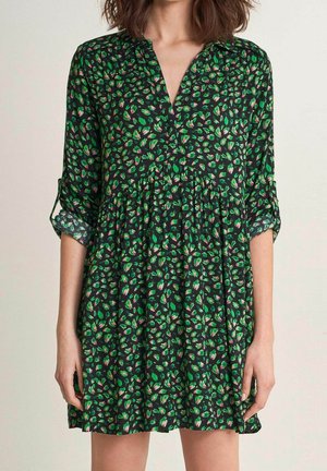 Kobieta ubrana w luźną czarną sukienkę z zielono-różowym kwiatowym wzorem, z podwiniętymi rękawami i dekoltem w serek.