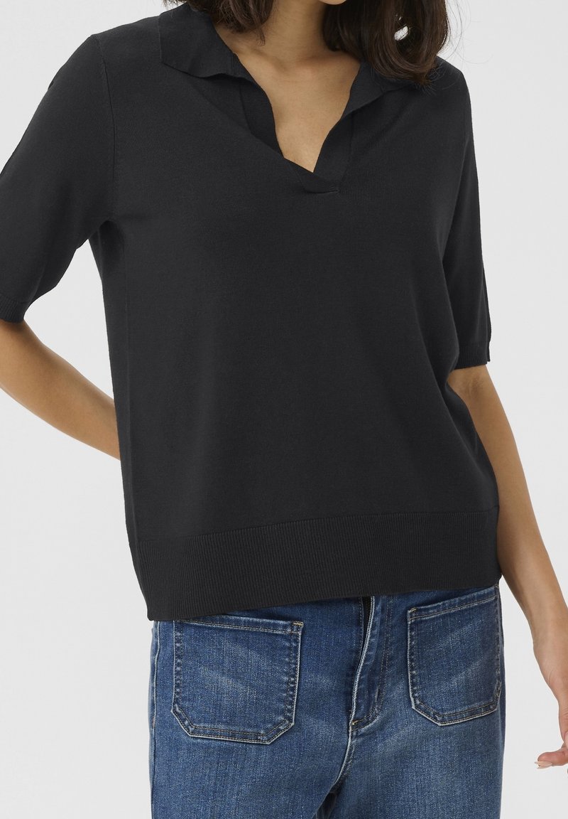 Polo noir à manches courtes avec col en V, poignets et ourlet inférieur côtelés. Porté avec un jean bleu clair, mettant en valeur une coupe décontractée.