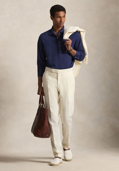 Άνδρας με μπλε μακρυμάνικο πόλο, κρεμ παντελόνι, λευκά εσπαντρίγιες, κρατώντας κρεμ σακάκι στον ώμο και καφέ δερμάτινη τσάντα ώμου στο χέρι.