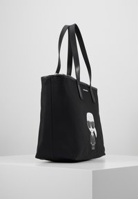 Svart nylon tote-väska med dubbla svarta handtag, med en vit grafik av en figur som bär solglasögon, dragkedjestängning, rektangulär form.