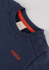 Camicia in cotone blu navy con un accento rosso sul colletto, dettaglio a rombi sulle spalle e logo "sigikid" stampato in rosso. Chiusura a pressione sul collo.