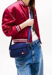 Giacca bomber in raso rosso con strisce bianche, abbinata a jeans azzurri chiari e una piccola borsa a tracolla blu navy con logo.