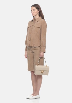 Donna in piedi che tiene una borsa beige, indossa una camicia color sabbia con bottoni e pantaloncini coordinati fino al ginocchio con scarpe bianche piatte, su sfondo bianco.