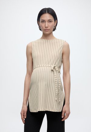 Gravid kvinne med kort mørkt hår iført ermeløs, kremfarget tunika med tynne vertikale striper og knyting på siden, kombinert med svarte bukser.