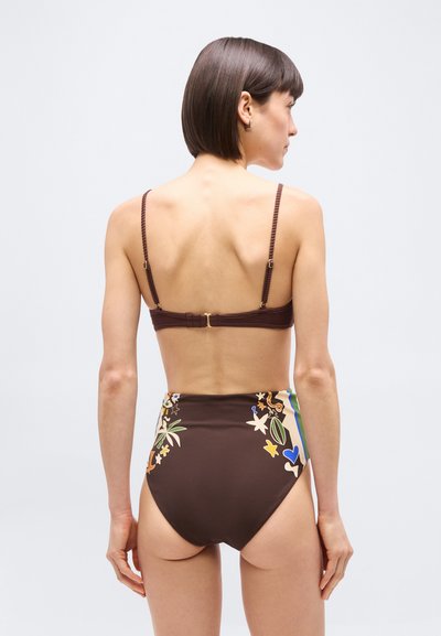 Γυναίκα με κοντά καστανά μαλλιά, φοράει καφέ ριγωτό σουτιέν και μαγιό σλιπ με φλοράλ σχέδιο και ψηλή μέση, γυρισμένη προς τα πίσω με φόντο ένα απλό σκηνικό.