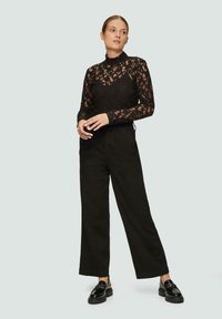 Schwarzes Spitzentop mit hohem Kragen, verziert mit floralen Mustern und langen Ärmeln, kombiniert mit hochtaillierten schwarzen, weiten Hosen und glänzenden Loafers.