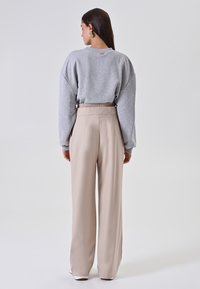 Donna con lunghi capelli scuri indossa una felpa grigia, pantaloni beige a vita alta e larghi, e scarpe bianche, in piedi davanti a uno sfondo bianco.