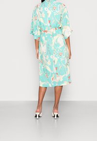 Luźna, turkusowa sukienka w kwiatowy wzór, z rękawami do połowy przedramienia i paskiem, w połączeniu z białymi sandałami na obcasie.