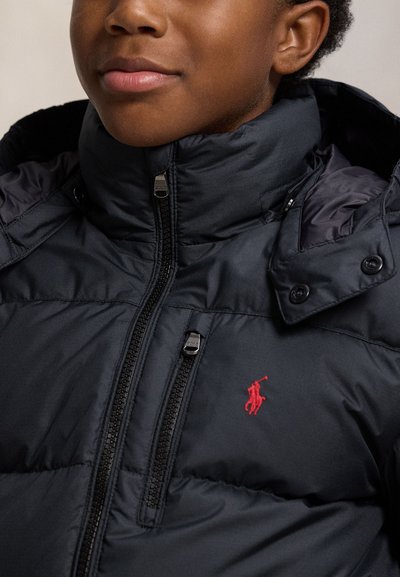 Dítě má na sobě černou prošívanou bundu s vysokým límcem zapnutou na zip, která má na hrudi malý červený vyšívaný znak.