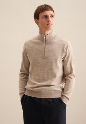 Beige Zip-Pullover aus weichem Strickmaterial mit langen Ärmeln, einem gerippten Kragen und schmalen Bündchen, getragen über dunklen Hosen.