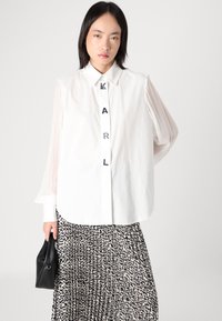 Chemise blanche à manches transparentes bouffantes, arborant le texte 'KARL' sur le devant. Associée à une jupe plissée noire et blanche à motif.