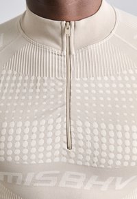 T-shirt de performance beige à manches longues avec un demi-zip, des motifs texturés et un tissu lisse avec des accents côtelés au niveau du col.