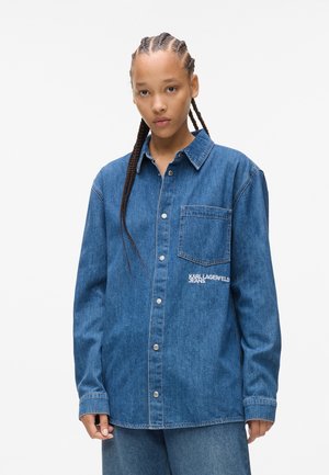 Modrá džínová košeľa s gombíkovým zapínaním, vreckom na prsiach a bielou výšivkou loga. Trochu voľnejší strih so rovnou líniou.