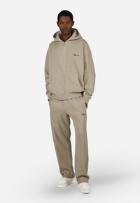 Beige ribbet hettegenser med glidelås, store frontlommer og logodetaljer, kombinert med matchende ribbet bukser og hvite joggesko.