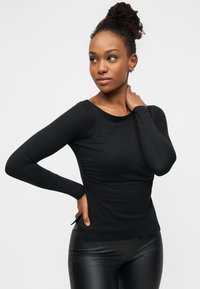 Εφαρμοστό μακρυμάνικο μαύρο τοπ από λείο, ελαστικό ύφασμα με στρογγυλή λαιμόκοψη και πλευρικές σχισμές, συνδυασμένο με γυαλιστερό μαύρο παντελόνι.