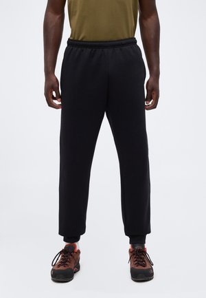 Personne portant un pantalon de survêtement noir et des chaussures de randonnée marron, debout sur un fond clair uni, les mains relâchées le long du corps.
