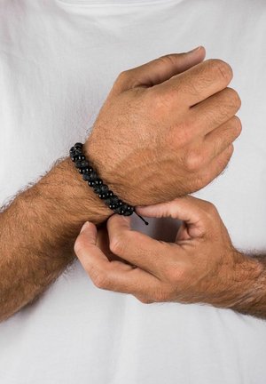 Melna pērlīšu aproce, kurā apvienotas gludas un teksturētas pērlītes, savilkta uz melna regulējama aukliņa, valkāta uz plaukstas ar gaišu ādas toni.