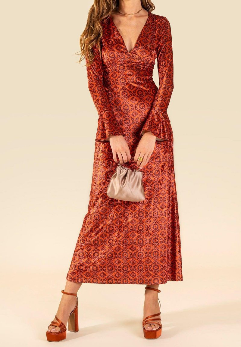Femme portant une longue robe rouge à motifs avec des manches évasées, tenant une petite pochette beige et portant des talons en plateforme marron.