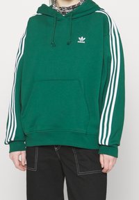 Zelená mikina s kapucí z měkkého materiálu, s bílými vertikálními pruhy na rukávech a přední kapsou. Logo Adidas na hrudi.