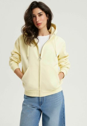 Kobieta o ciemnych falowanych włosach, ubrana w bladożółtą bluzę z kapturem zapinaną na zamek, na białym topie i jasnoniebieskich dżinsach, stoi z rękami w kieszeniach.