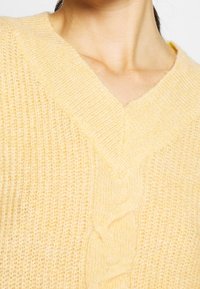 Pull jaune tricoté avec un col en V, présentant un motif torsadé sur le devant, une texture douce et des poignets côtelés.