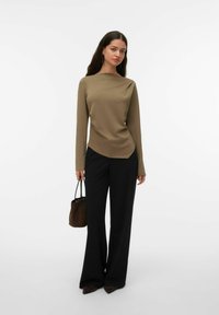 Langermet olivengrønn topp med asymmetrisk kant, kombinert med høytlivede svarte bukser. Tilbehør inkluderer en liten brun veske.