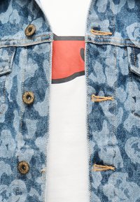 Τζιν μπουφάν με μπλε λουλουδένιο ντεγκραντέ και μπρούτζινα κουμπιά πάνω από λευκό πουκάμισο με κόκκινο και μαύρο αφηρημένο σχέδιο.