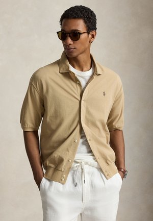 Mann trägt Schildpatt-Sonnenbrille, beiges kurzärmeliges Hemd mit Knöpfen über weißem T-Shirt, weiße Hose mit Kordelzug, Hände in den Taschen.