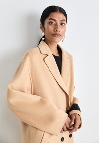 Cappotto in lana beige con colletto a revers, design doppiopetto e abbellimenti in bottone nero. La modella indossa un top nero e orecchini dorati.