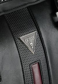 Zwart lederen oppervlak met een gestructureerde zwarte band en een driehoekig metalen label met het woord "GUESS" en een vraagteken, geaccentueerd met rood.