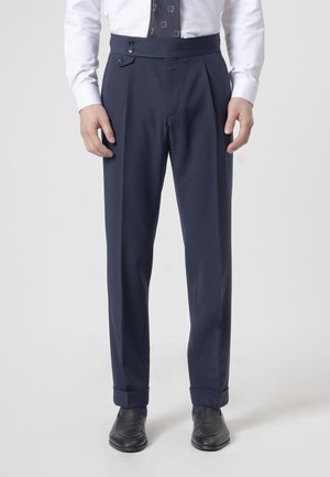 Hombre con pantalones de traje azul marino de talle alto, camisa blanca de vestir, corbata estampada y zapatos de vestir negros, de pie sobre un fondo liso.