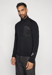 Jachetă neagră cu fermoar, având un guler înalt, o buzunar texturat pe piept și un detaliu cu logo aproape de margine. Fabricată dintr-un material neted și elastic.