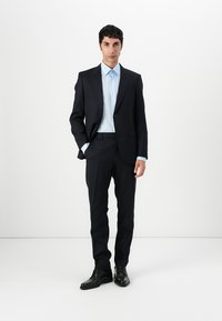 Dunkelblauer Anzug mit taillierter Passform, kombiniert mit einem hellblauen Hemd. Verfügt über ein Ein-Knopf-Jacket, glattes Material und glänzende schwarze Schuhe.