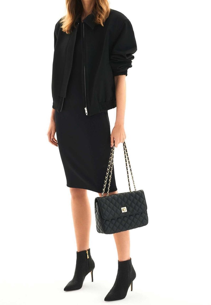 Femme portant une robe noire et une veste, tenant un sac à main noir matelassé avec des chaînes dorées en bandoulière, et portant des bottines noires à talons hauts.