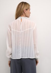 Hvid transparent bluse med vertikale striber, en perlekant og pufærmer. Har en knaplukning bagpå og elastiske ærmer.