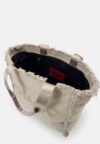 Beige Einkaufstasche mit fransigem Rand, schwarzem Innenfutter, zwei Griffen und einem roten Innenetikett mit "HUGO"-Branding.