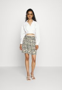 Camisa branca cropped com mangas longas, saia estampada de flores em cores escuras e sandálias brancas de salto. Os acessórios incluem óculos de sol amarelos.