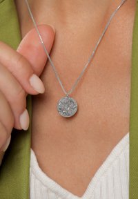 Ezüst medálos nyaklánc, strukturált, kerek medállal, finom láncra függesztve. Világos színű felső és zöld dzseki mellett viselve.
