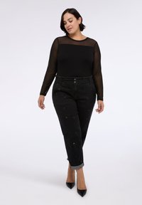 Donna che indossa un top lungo a maniche sheer nera, pantaloni neri con maniche decorate e tacchi a punta neri, che cammina con gli occhi chiusi su uno sfondo bianco.