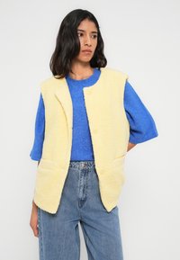 Gilet jaune texturé avec fermeture à boutons et poches frontales, porté sur un pull à manches courtes bleu et un jean bleu taille haute.