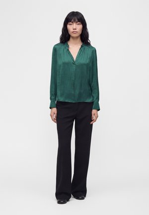 Femme portant un chemisier vert brillant à manches longues et un pantalon noir à jambes larges, debout devant un fond clair uni.