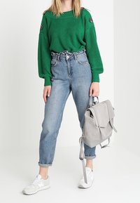 Donna che indossa un maglione verde a maglia e jeans blu a vita alta, con uno zaino grigio chiaro in mano, in piedi con sneakers bianche.