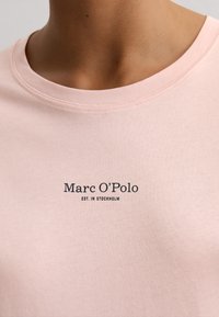 Close-up van een persoon die een lichtroze t-shirt draagt met "Marc O'Polo EST. IN STOCKHOLM" in zwarte letters op de borst gedrukt.